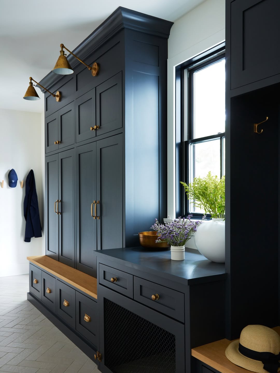 Dark blue built-in cabinets offer stylish storage solutions, paired with brass hardware, a wooden bench, and a counter topped with a white vase of flowers beside the window.