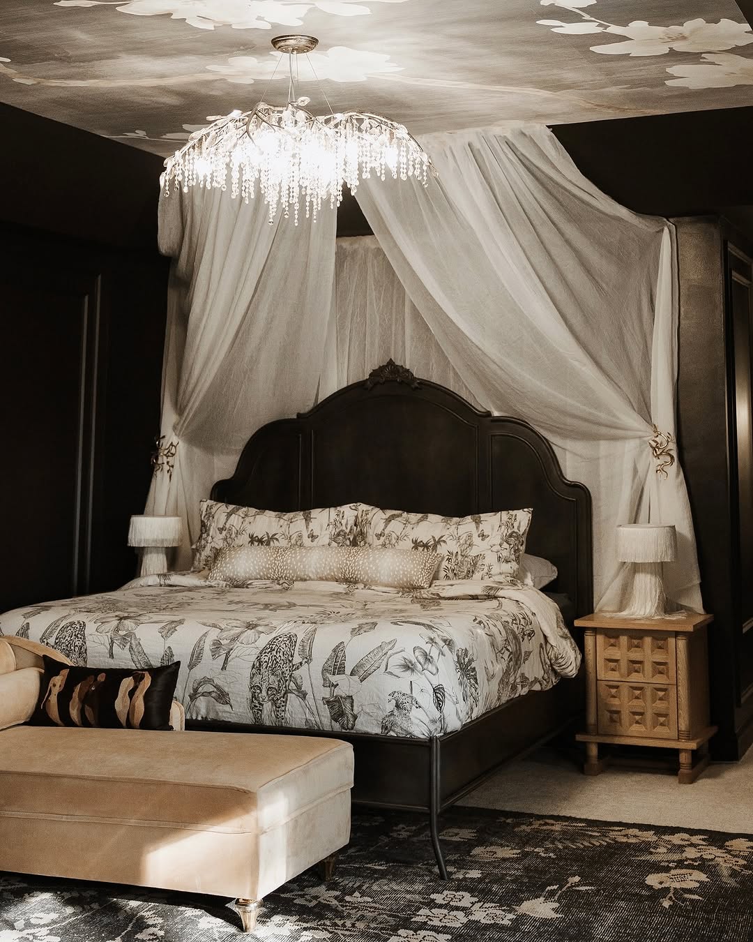 A dark feminine bedroom featuring a dark wood bed frame, white floral bedding, sheer canopy drapes, two small bedside tables with lamps, a patterned rug, and a beige upholstered bench.