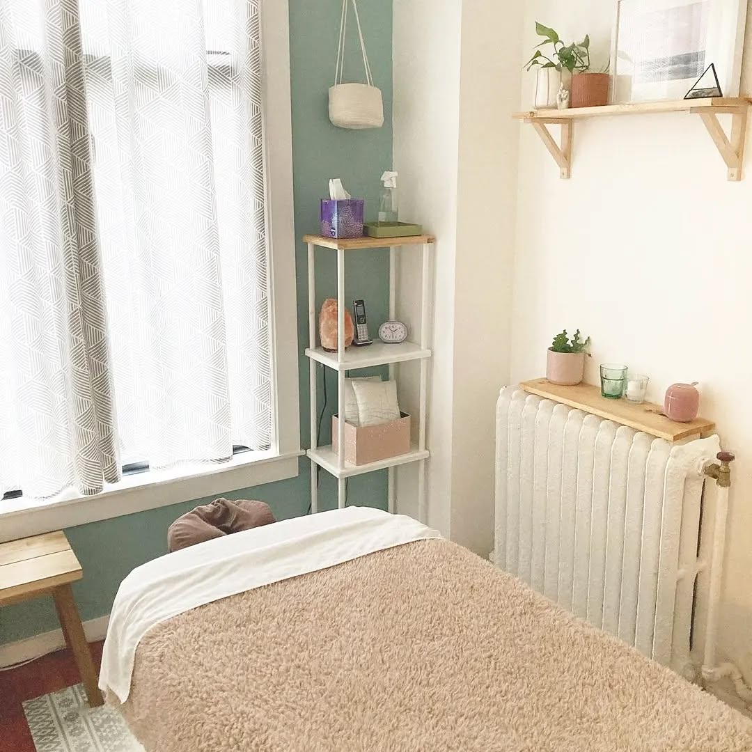 A cozy massage room design features a massage table, a shelving unit holding tissues and decor, a radiator, and a window adorned with patterned curtains.