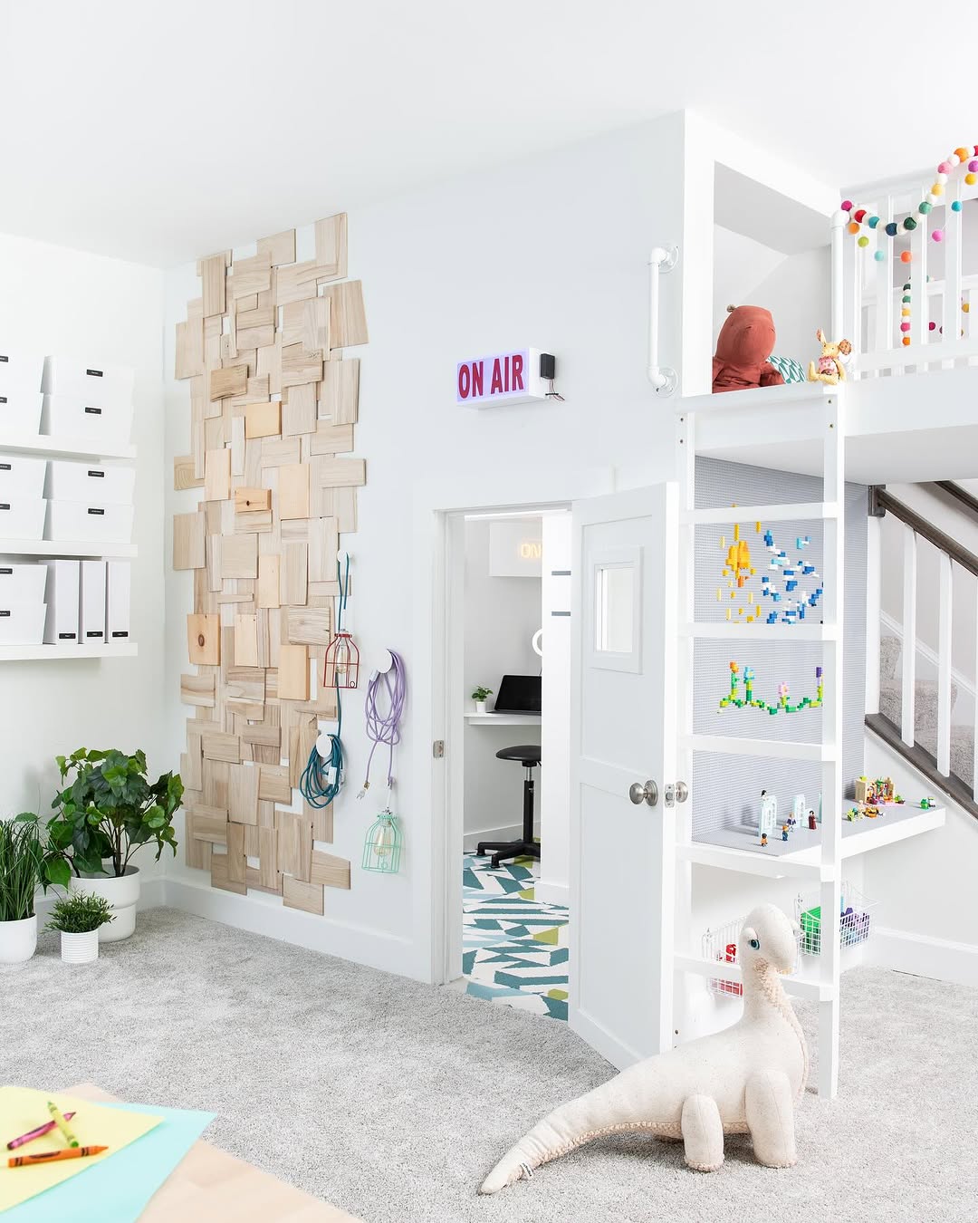 A modern, bright under stair playroom features a loft bed, wall-mounted storage boxes, a geometric wood accent wall, plants, toys, and an