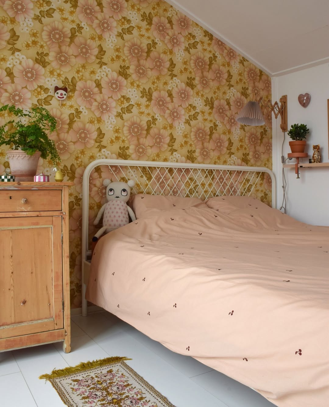 A neatly made bed with peach bedding sits against retro floral wallpaper. Plush dolls, plants, and a wooden cabinet add charm—perfect inspiration for chic Cherry Bedroom Ideas in a cozy space.