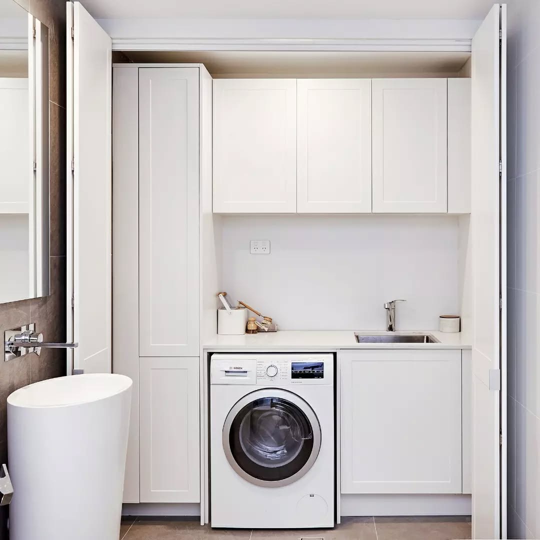 Compact laundry area with white laundry cabinets, a front-load washing machine, sink, and countertop, all neatly built into a recessed space with open double doors for smart laundry room storage.