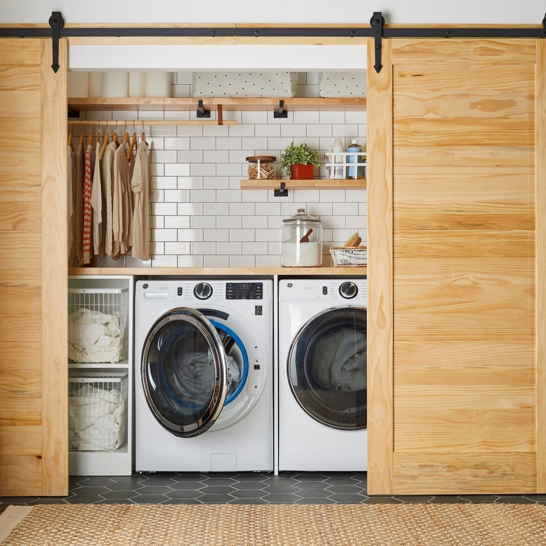 A Boho laundry room featuring a front-loading washer and dryer, open shelving with jars and baskets, hanging clothes, stylish laundry room decor, and rustic sliding wooden barn doors.