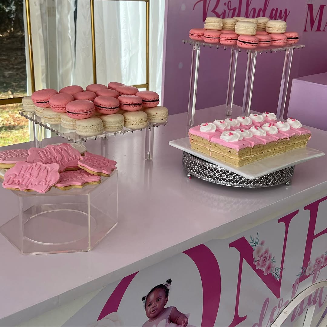 A dessert table featuring memorable desserts like pink and white macarons, rectangular cakes with pink and white frosting, and pink iced birthday cookies, all set in front of a festive birthday-themed backdrop.
