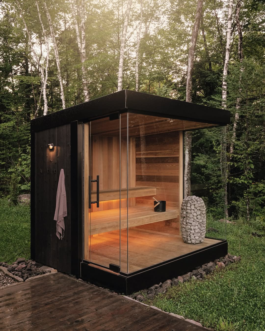 This small modern sauna with glass walls and a wooden interior sits in a forested area—a stunning example of outdoor sauna ideas, with a towel on the exterior wall and stones stacked by the heater inside.