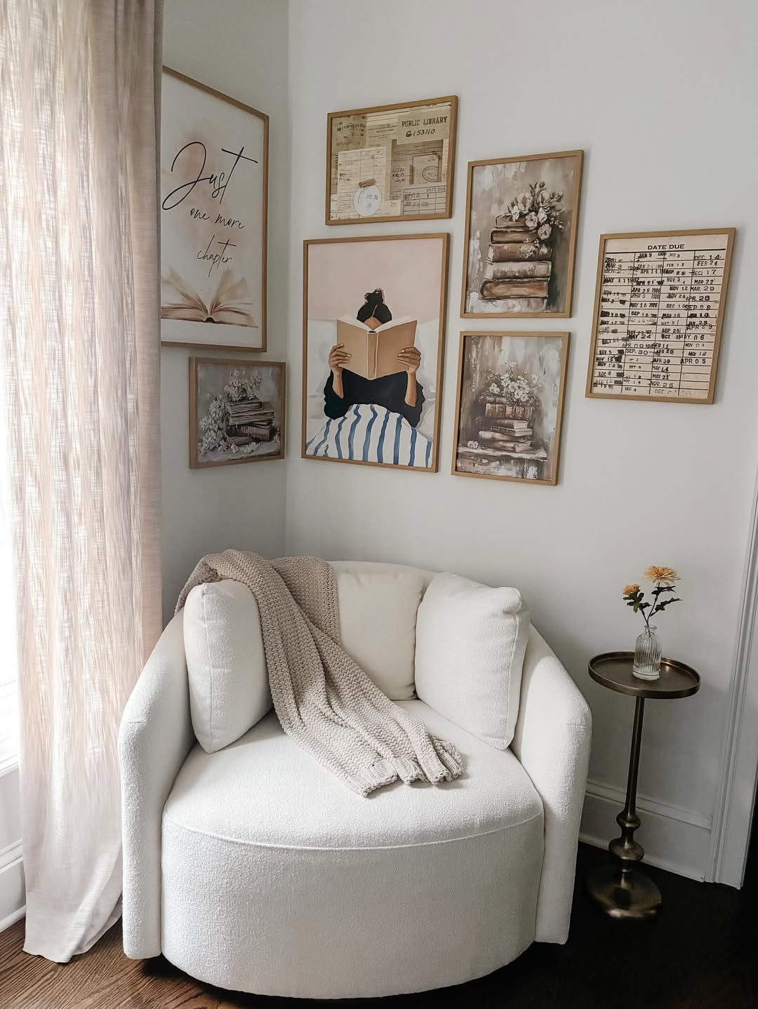 A white, rounded armchair with a beige throw sits in a corner by a window, creating a cozy reading nook next to a small side table with flowers; framed art and photos decorate the wall above.