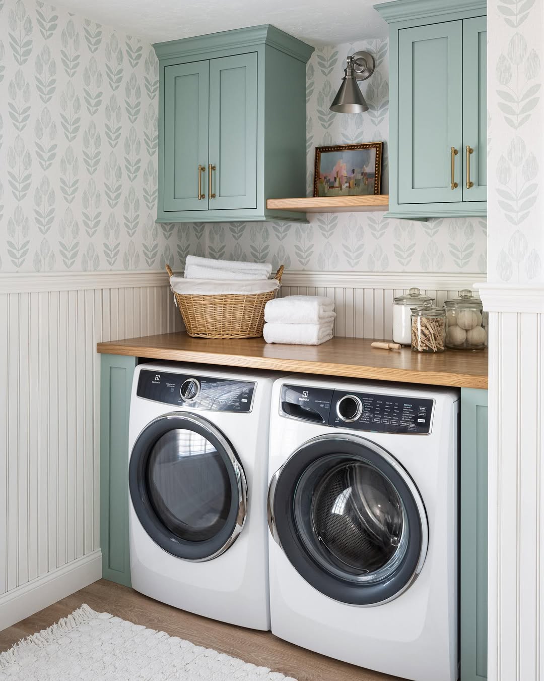 A small laundry room features a washer and dryer tucked under a wooden countertop. Green cabinets hover above, while a wicker basket with towels, a photo, and glass jars grace the shelf. Leaf-patterned wallpaper adorns the walls, adding charm to this compact space.