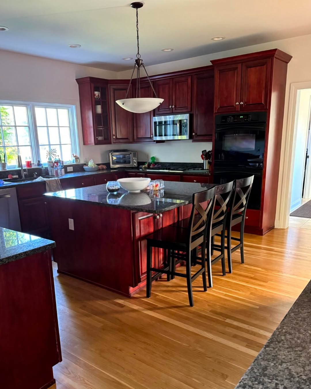 Modern kitchen with dark wood cabinets, black countertops, and a center island with three chairs, showcasing stunning kitchen colors. Built-in appliances and a large window let in natural light, reflecting the latest kitchen color trends 2025.