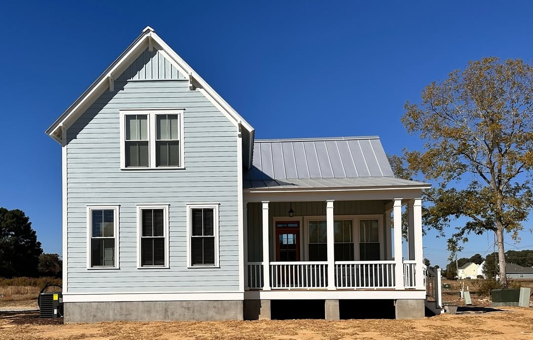 A light blue, two-story house with a covered front porch, white trim, and metal roof showcases charming Coastal cottage exterior design ideas, set on a dirt lot with a tree and clear blue sky in the background.