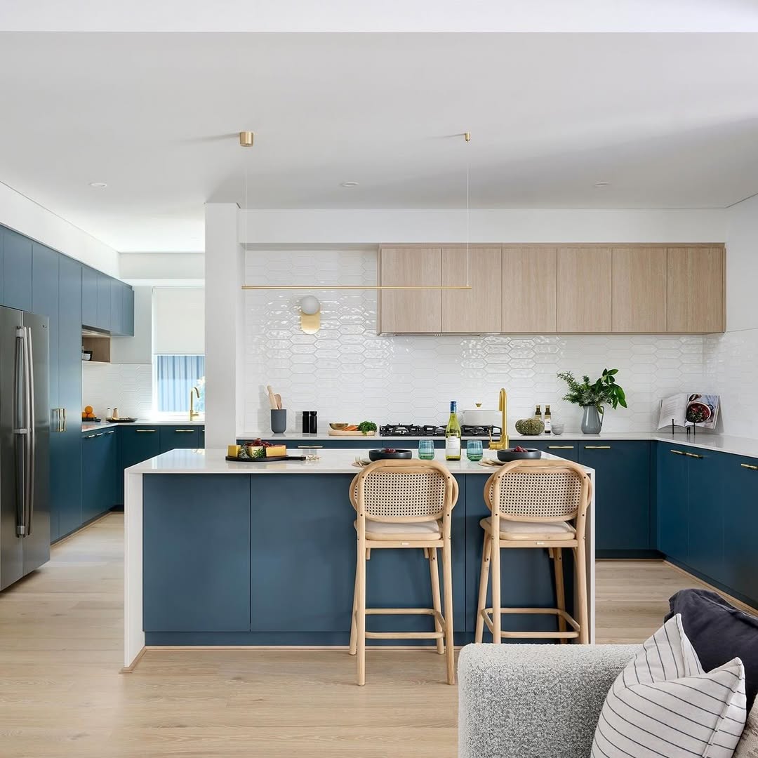 Modern kitchen with stunning blue lower cabinets, light wood uppers, a white tile backsplash, and two wooden bar stools—a perfect showcase for 2025 kitchen color ideas. Various kitchen items add charm to the stylish counter.