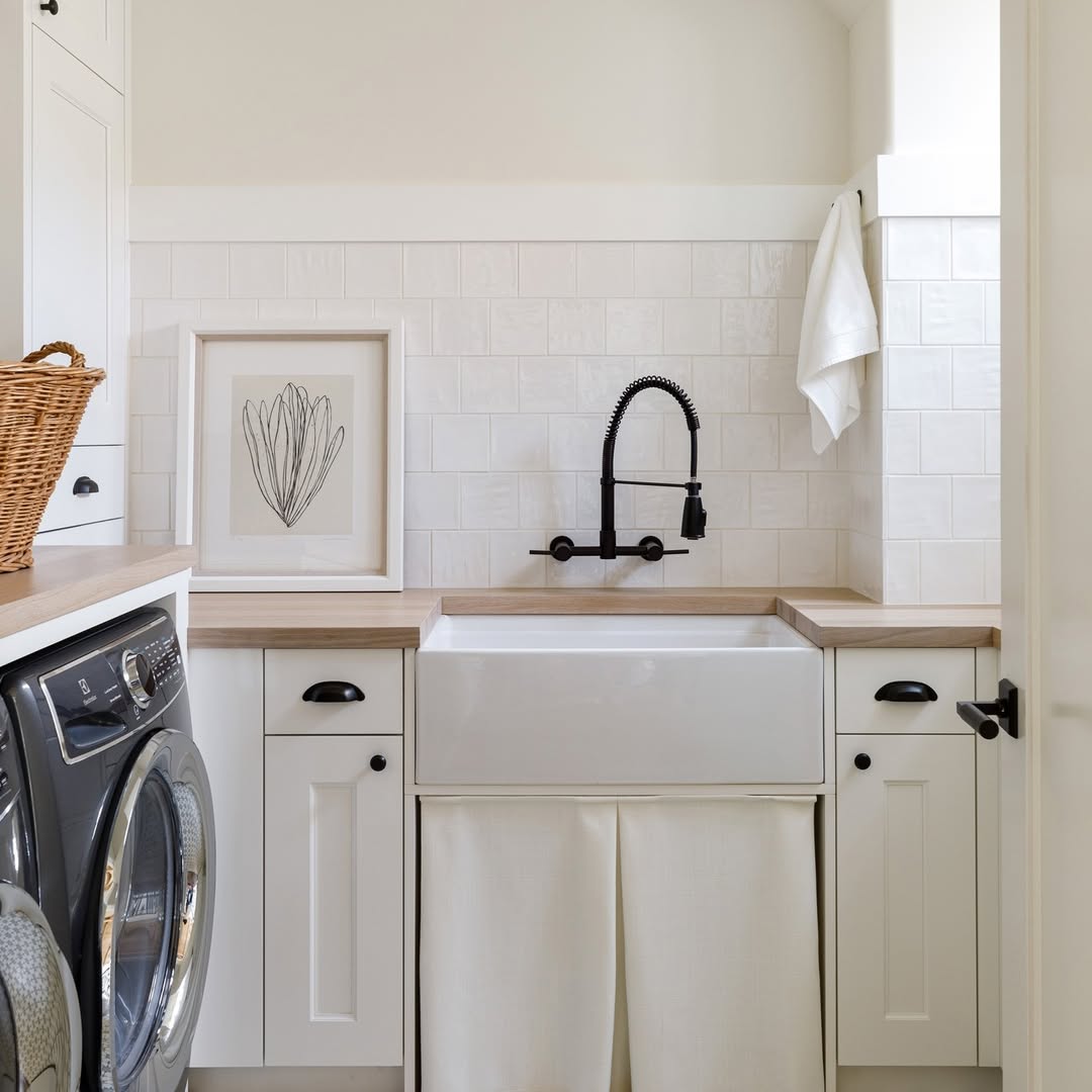 Laundry room featuring white laundry room cabinets, a front-loading washer, farmhouse sink with black faucet, wood countertops, a framed print, and a wicker basket.