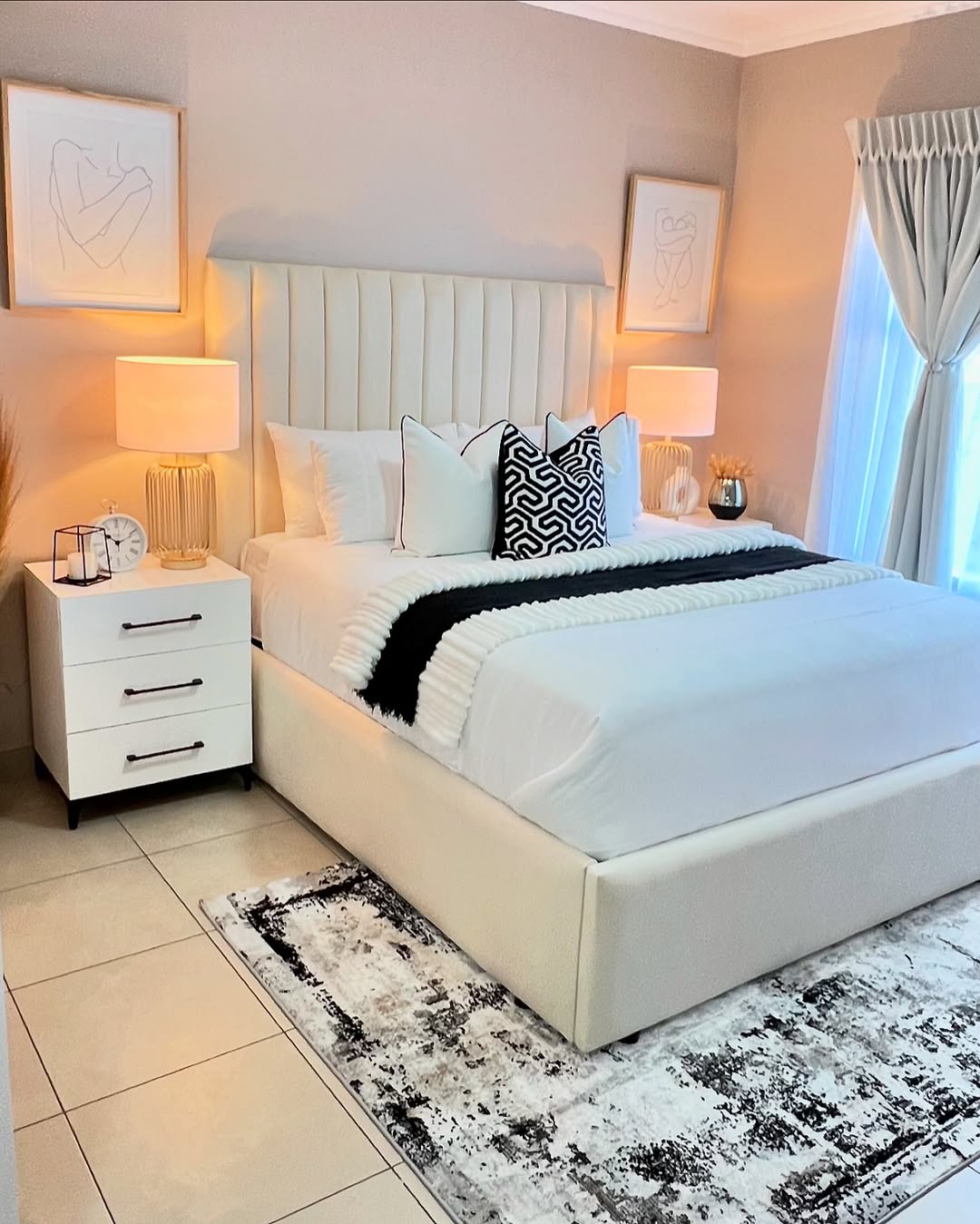 A modern bedroom design with a white upholstered bed, patterned rug, two nightstands with lamps, and minimalistic wall art. Light streams in through a window with sheer curtains, offering inspiring bedroom decor ideas.