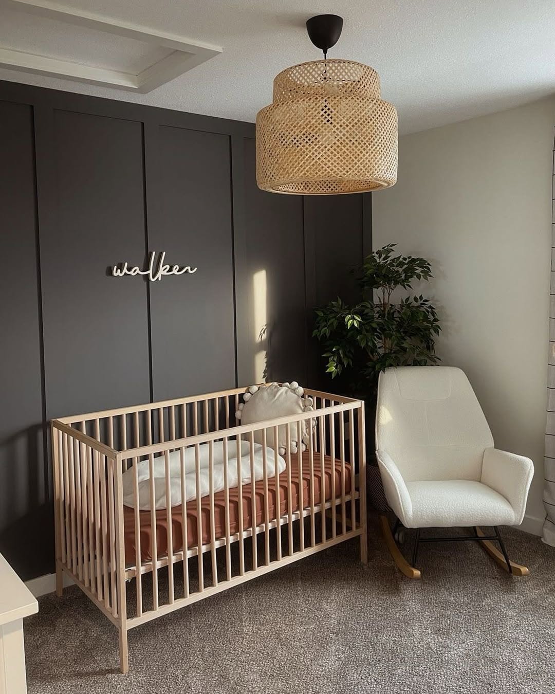 A modern nursery with a wooden crib, white rocking chair, faux plant, woven ceiling light, and “walker” name sign on a dark paneled accent wall—perfect inspiration for first-time moms seeking baby boy nursery ideas in 2025.