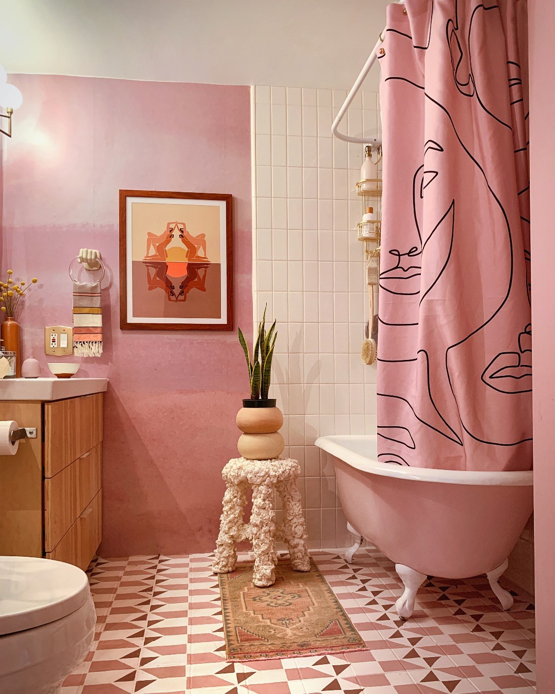 Charming pink bathroom with pink walls and floor tiles, a clawfoot tub with a matching shower curtain, wooden vanity, framed artwork, and a potted plant on a decorative stool.