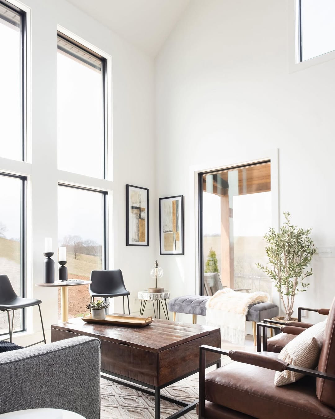 A modern chic living room with tall windows, neutral-toned furniture, dark brown sofas, two black chairs, framed artwork, a potted plant, and a wooden coffee table on a patterned rug.