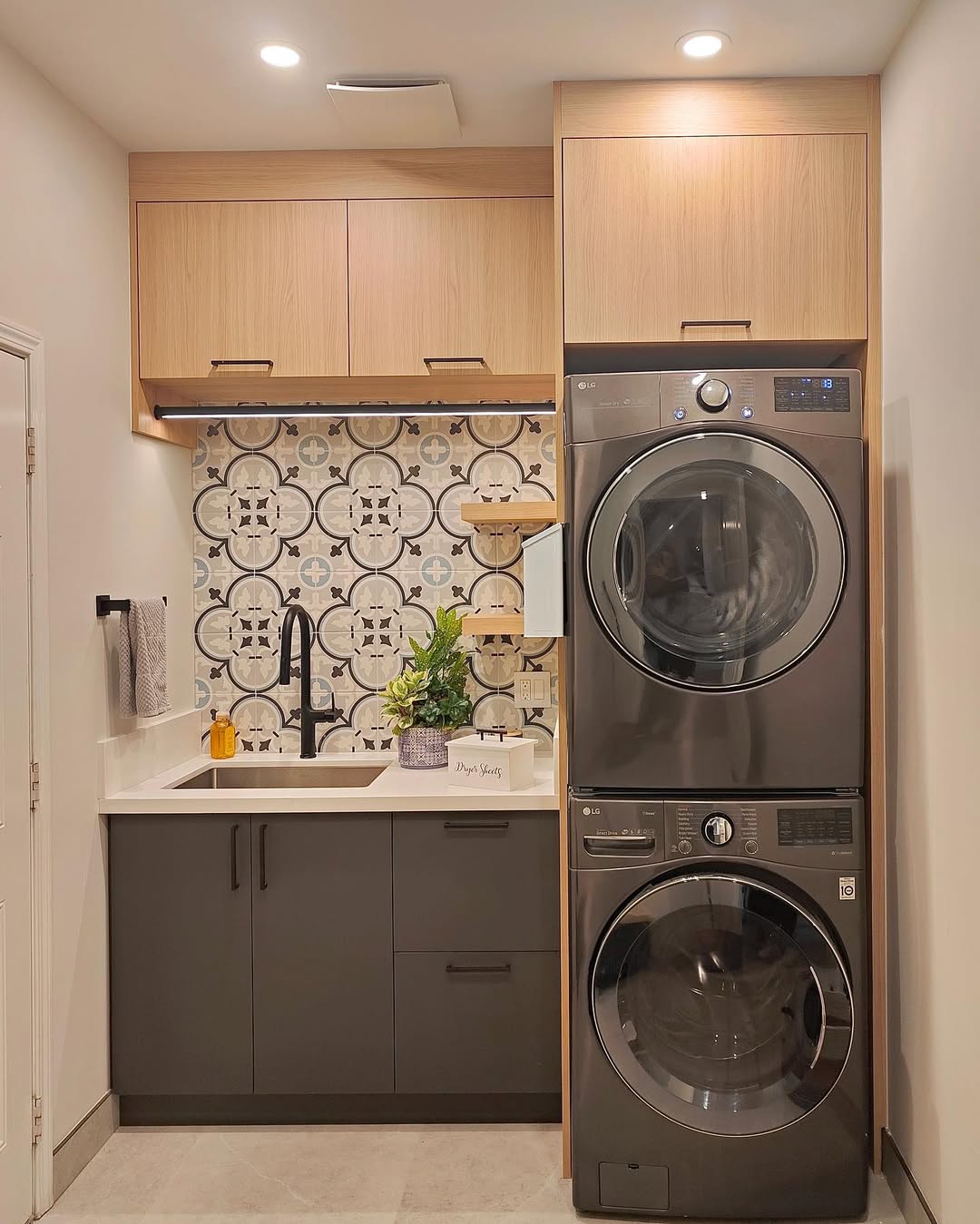 Modern stacked laundry room with washer and dryer, patterned tile backsplash, sink, gray cabinets, upper wood cabinets, and wall-mounted shelves with decor.