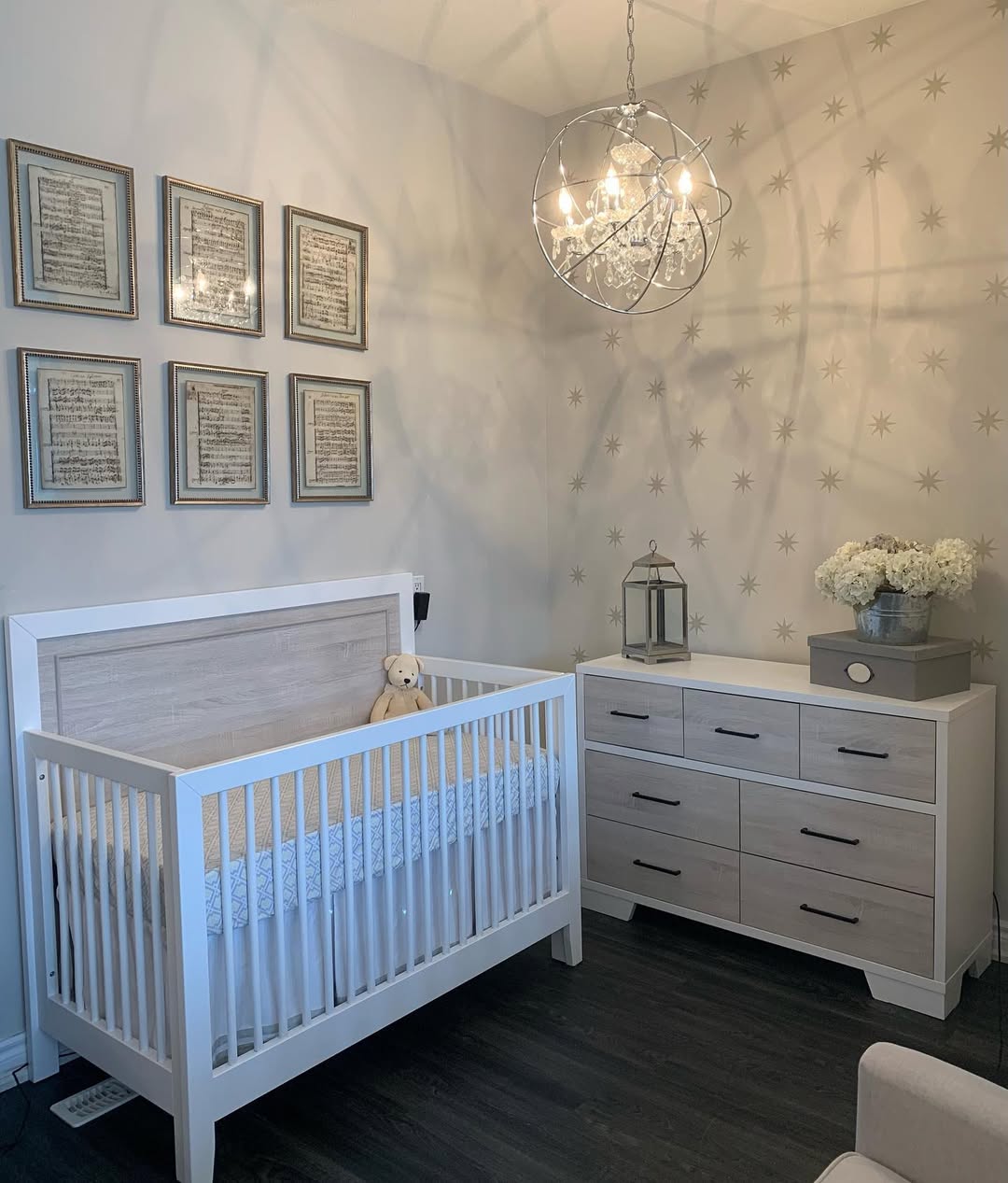 A modern nursery with a white crib, a teddy bear, chic nursery ideas like a sleek dresser with flowers, framed art on the wall, and a chandelier casting star-like shadows—perfect inspiration for first-time moms.