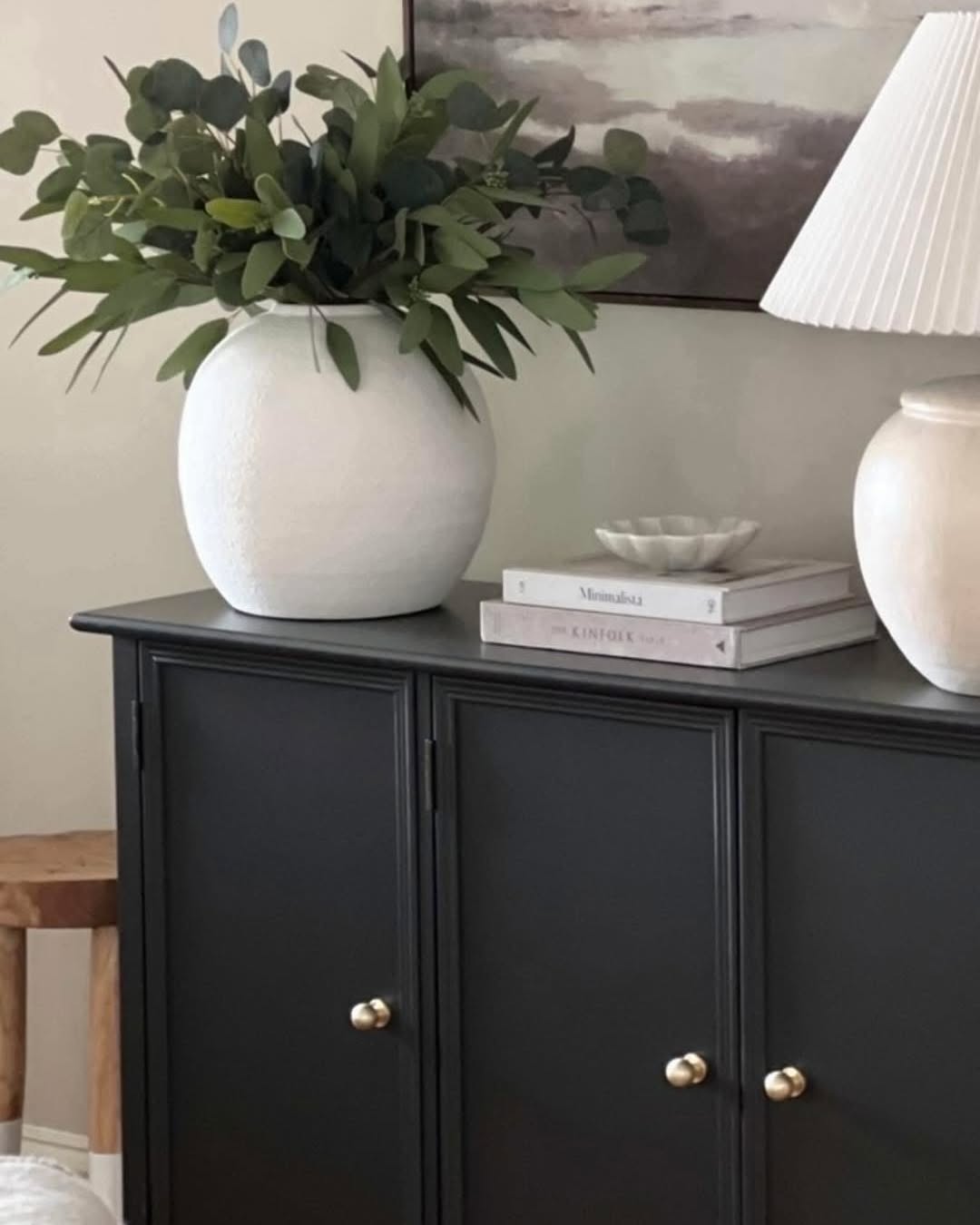 A white vase decor with green foliage, a table lamp, and stacked books sit on a dark cabinet against a light wall.
