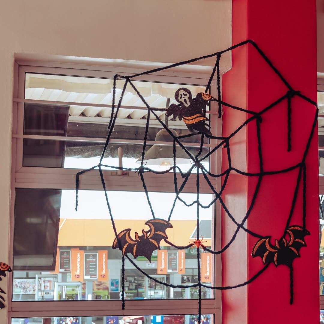 A Halloween decoration featuring a large black spider web with a ghost and two bats makes for creative window decor, attached to a red wall near a window.