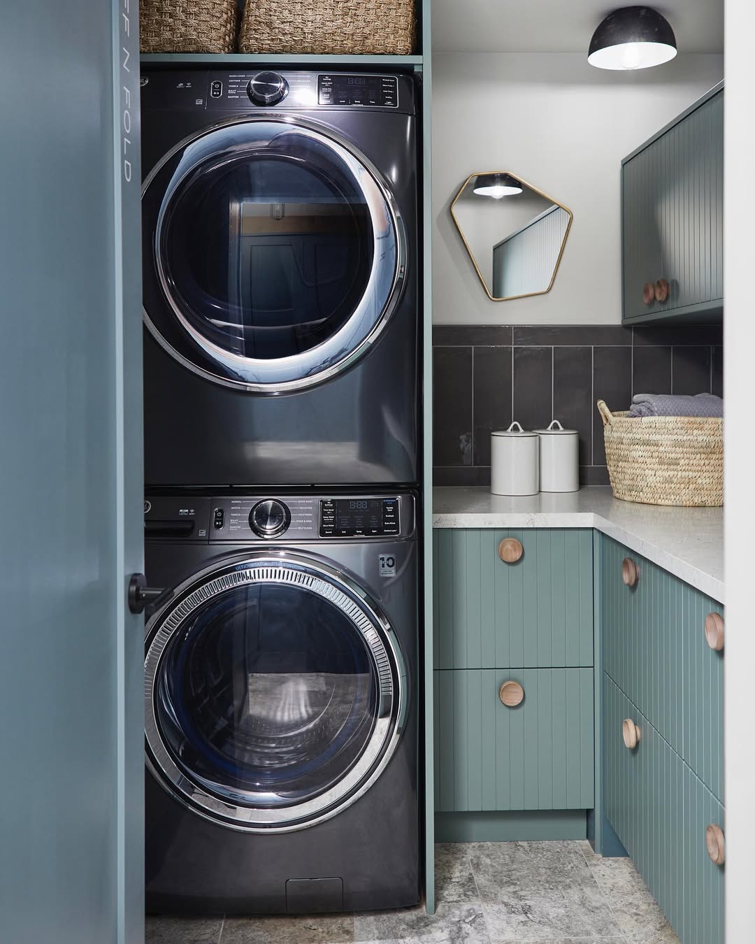 A modern stacked laundry room with teal cabinets, a countertop, a woven basket, wall tiles, a small mirror, and overhead lighting creates a stylish and functional space.
