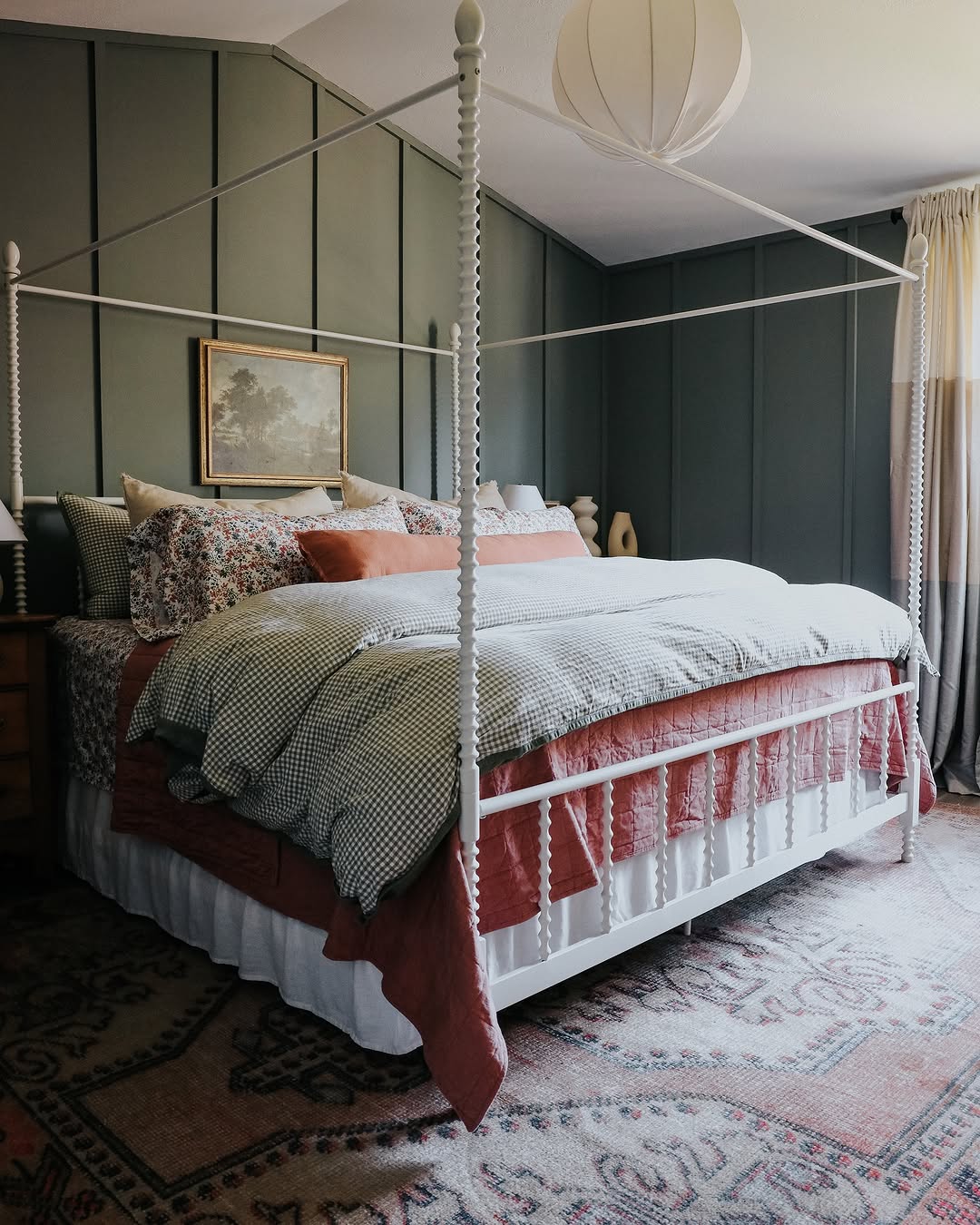 A neatly made four-poster bed with patterned bedding and pillows is centered in a dark feminine bedroom, featuring deep green paneled walls, a rug, and a hanging light fixture.