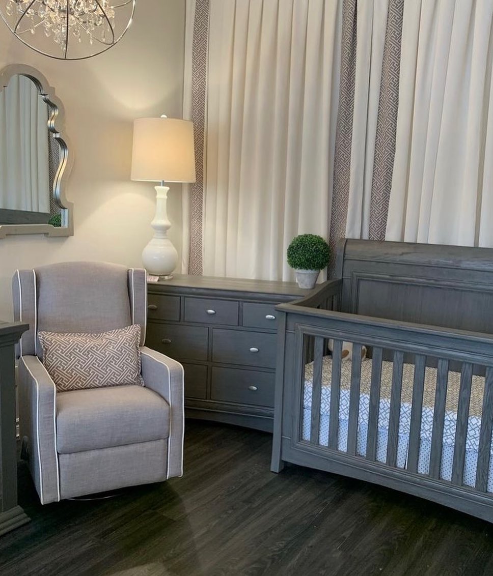 A gray nursery offers chic nursery ideas for a first-time mom, featuring a crib, armchair, dresser, table lamp, round mirror, decorative plant, and floor-to-ceiling curtains.