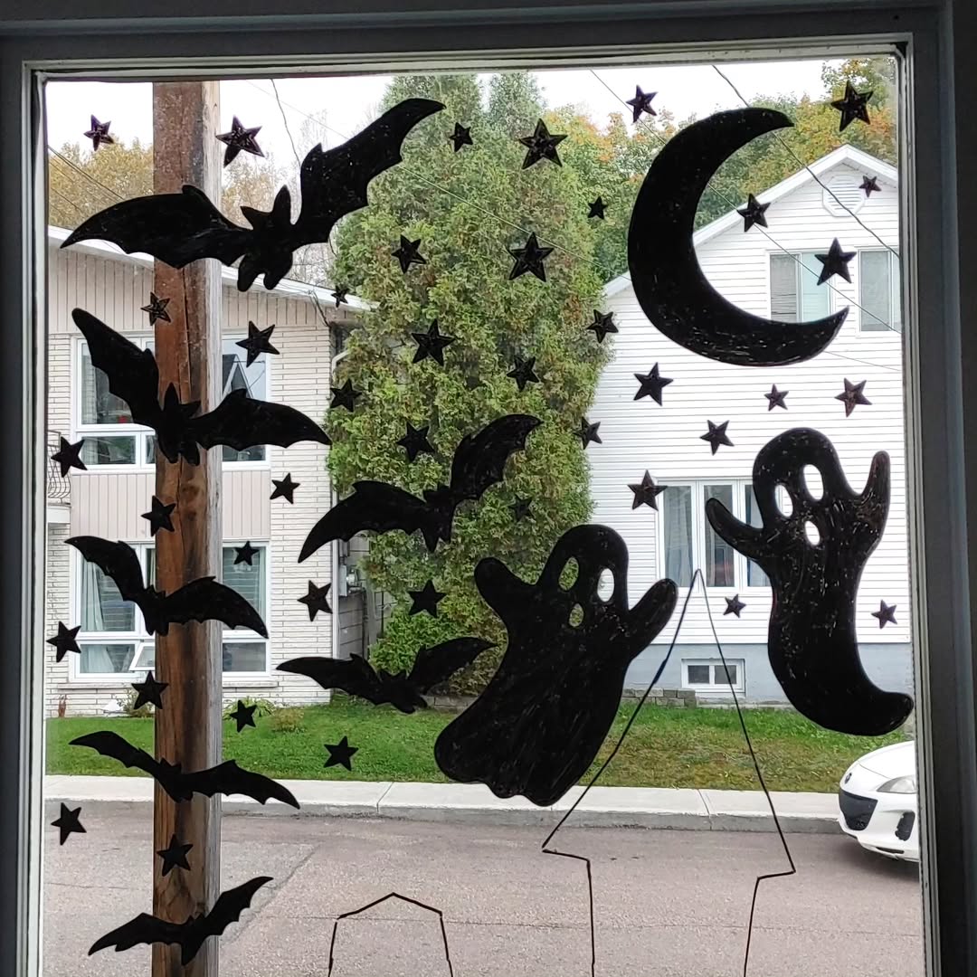 Paper cutouts of bats, ghosts, stars, and a crescent moon are taped to a window in one of the most charming Halloween window decoration ideas, with houses and trees visible outside.