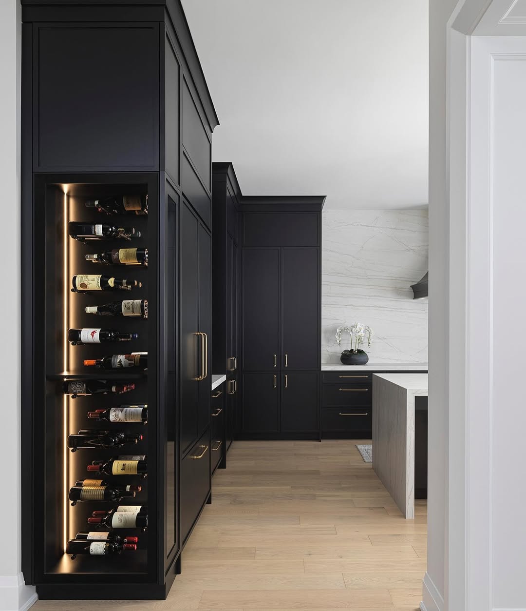 Modern kitchen with black cabinetry, light wood floors, and a built-in, illuminated wine rack—a stylish wine storage solution filled with assorted bottles. Marble backsplash and a white kitchen island complete the look.
