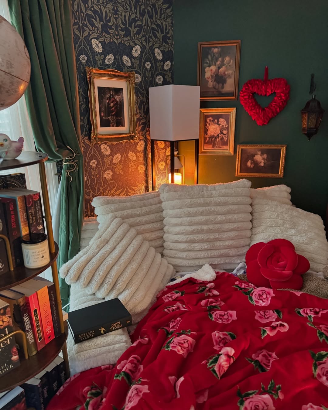 Cozy Reading Nook with a white sofa, fluffy pillows, red floral blanket, books, ornate art, a heart-shaped decoration, and warm lighting by a window with green curtains—perfect inspiration for your Book Nook Ideas collection.