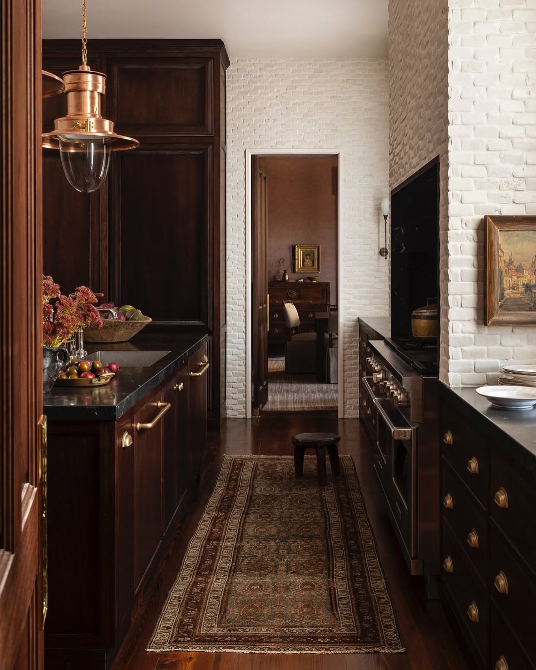 A narrow kitchen with dark wood cabinets, brass hardware, a patterned rug, and white brick walls showcases stunning kitchen colors while leading into another room through an open doorway.