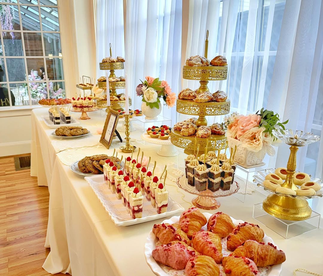A dessert table set with various party desserts—pastries, cakes, cookies, and parfaits—arranged on white tablecloths and gold stands near large windows with curtains offers delightful dessert table ideas for any celebration.