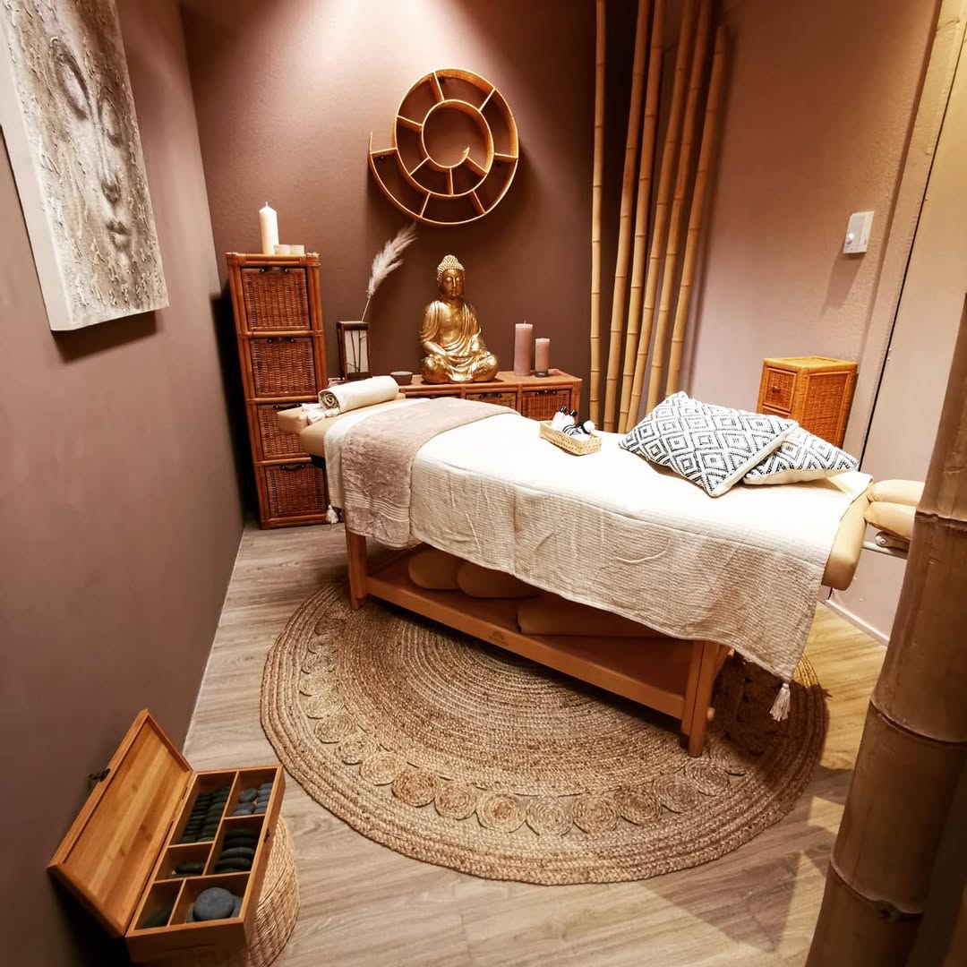 A massage table with neutral linens sits in a softly lit massage room decorated with bamboo, wicker furniture, candles, a Buddha statue, and a round woven rug—perfect for inspiring new massage room ideas.