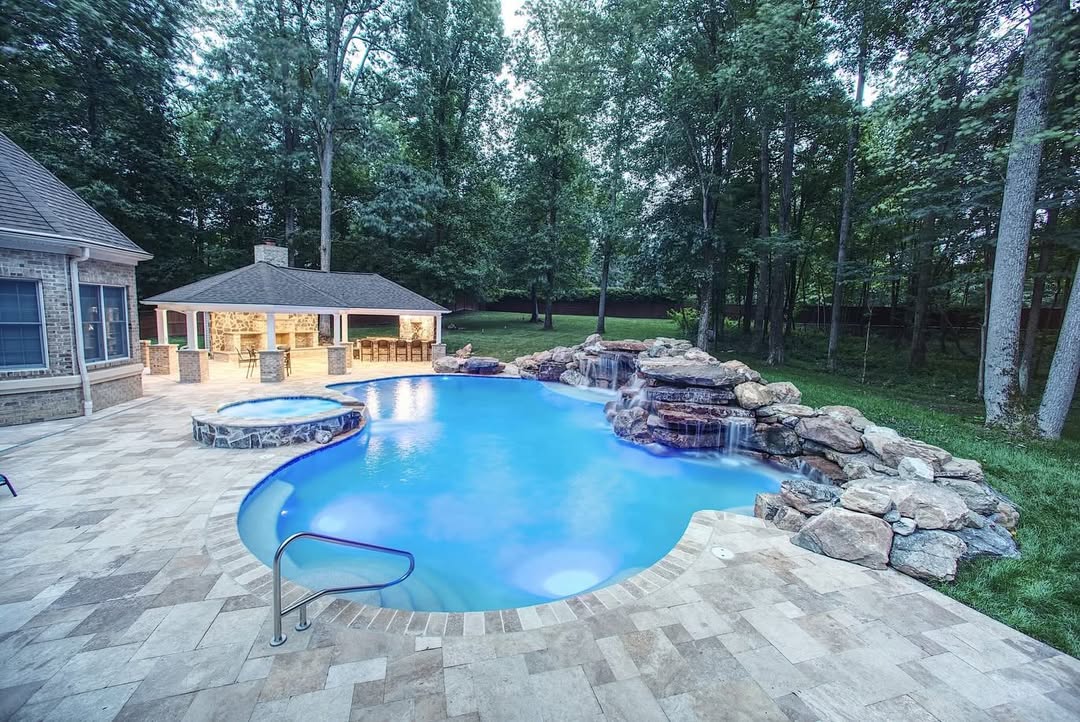 Outdoor swimming pool with stone waterfall feature, adjacent hot tub, and pool house—surrounded by trees and a stone patio—offers inspiring backyard pool ideas for your perfect retreat.