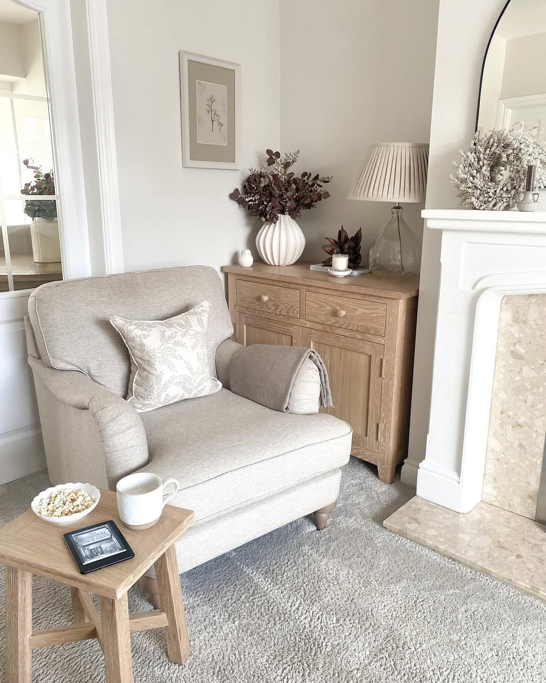 A beige armchair with a cushion sits beside a wooden side table holding a cup, a bowl of popcorn, an e-reader, and a charming vase decoration in a neutral-toned, cozy living room.