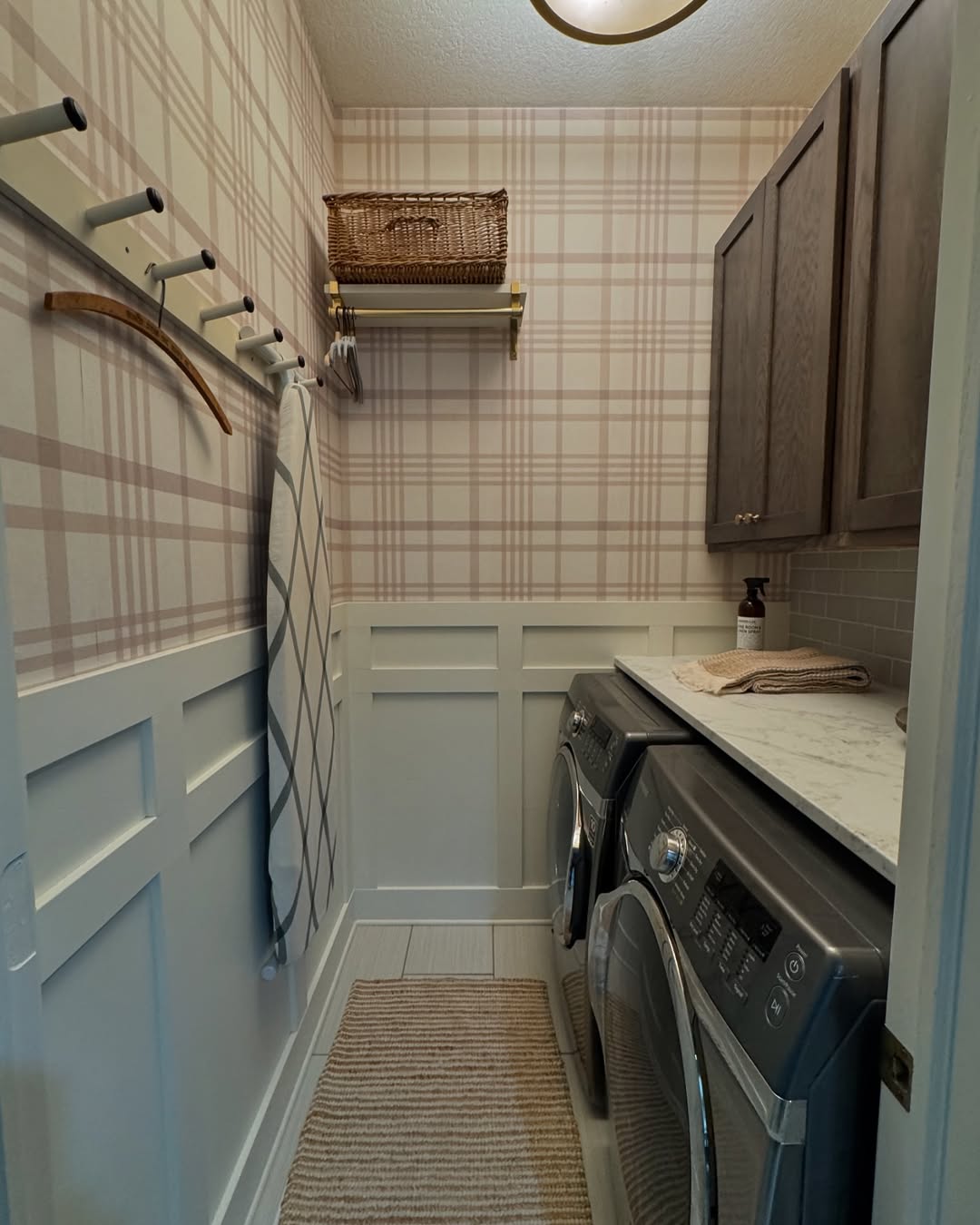 A small laundry room with plaid wallpaper features a clever laundry room design: washer and dryer, wall hooks holding a drying rack and towel, upper cabinets, a shelf with a basket, and a striped rug on the floor.