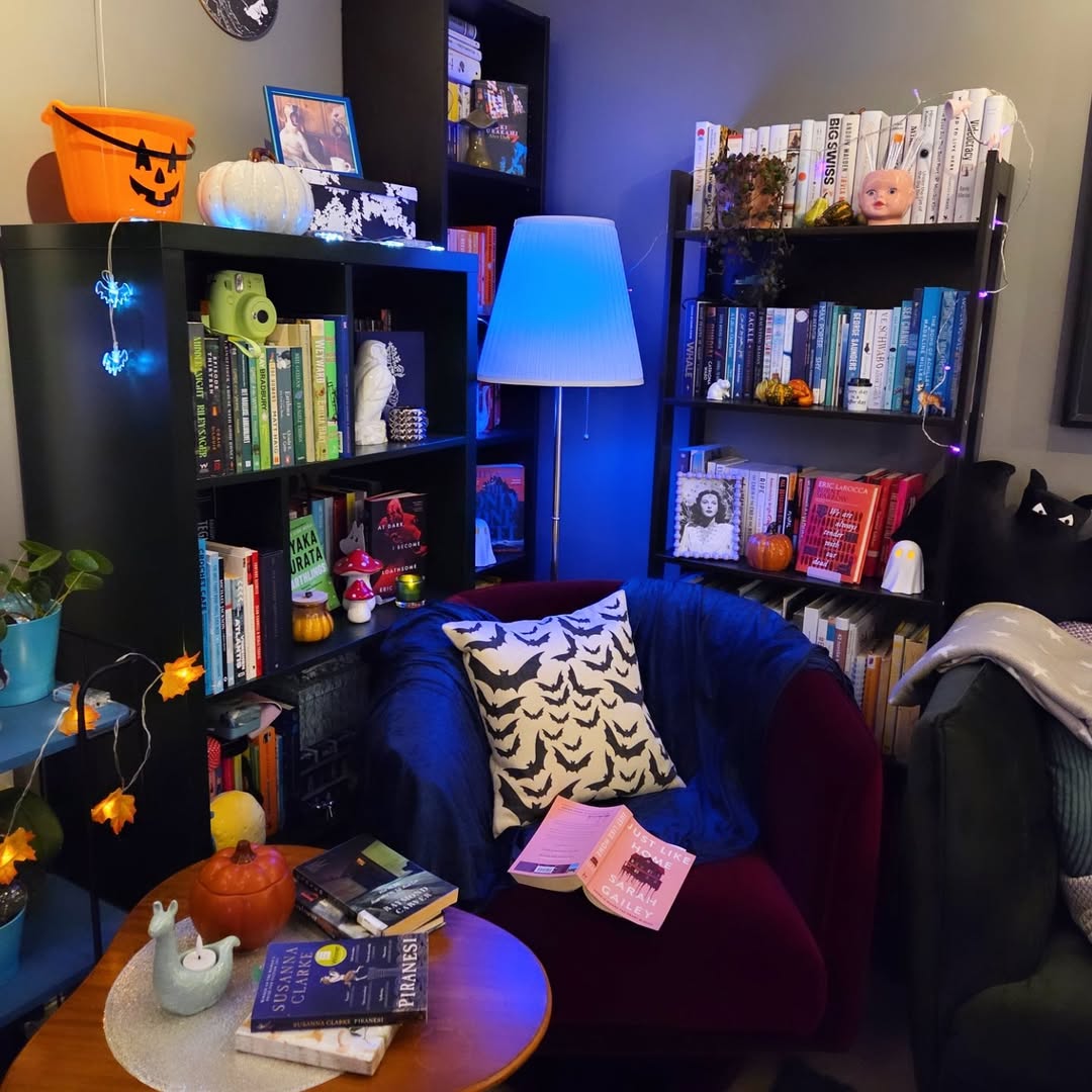 A cozy reading nook with bookshelves, a round table with books and a pumpkin, a lamp with blue light, plus Halloween decorations like string lights and a jack-o