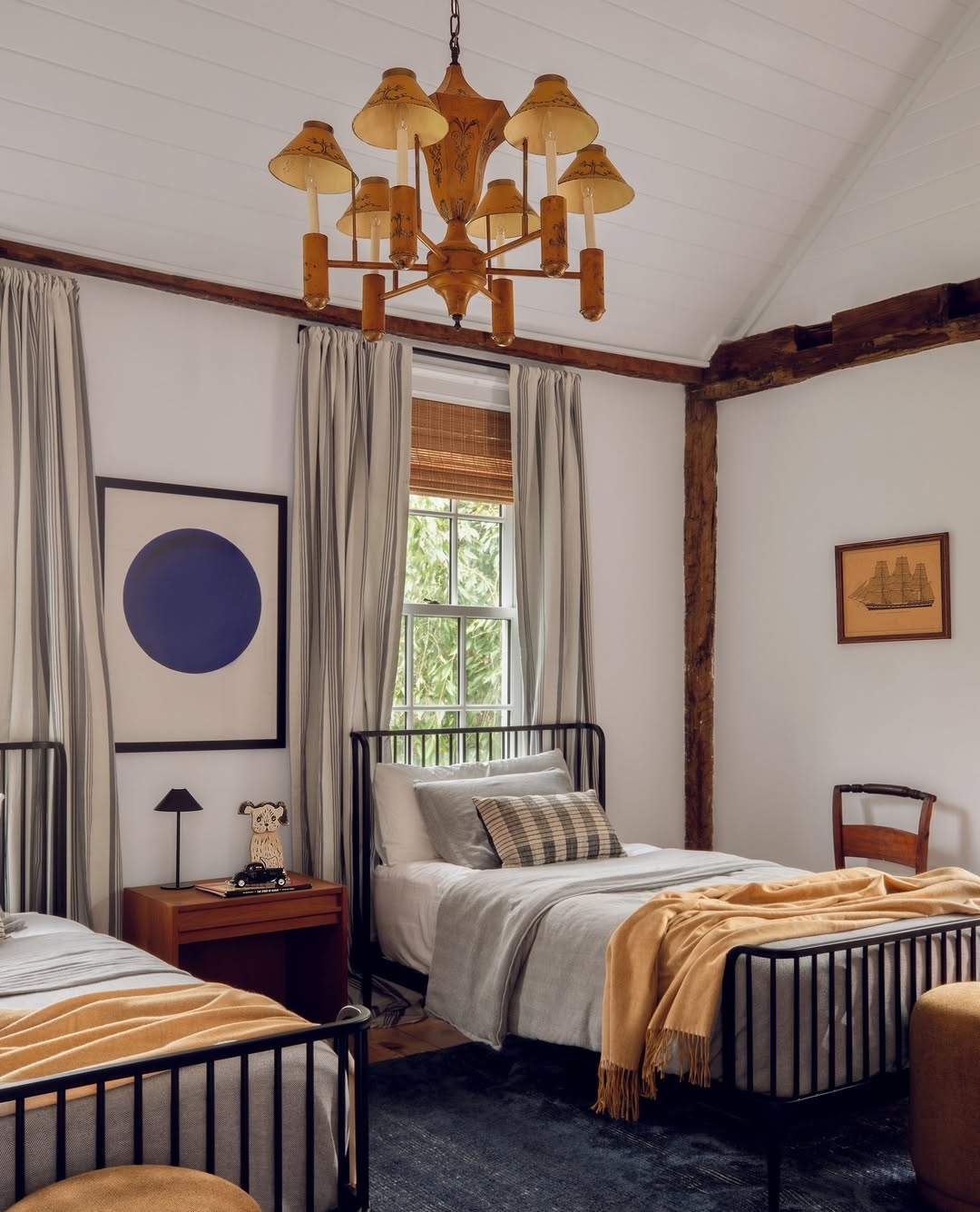 A cozy rustic bedroom with two metal-frame twin beds, neutral bedding, a wooden chandelier, artwork on the walls, and a window with gray curtains letting in natural light—perfect for those seeking rustic bedroom ideas.