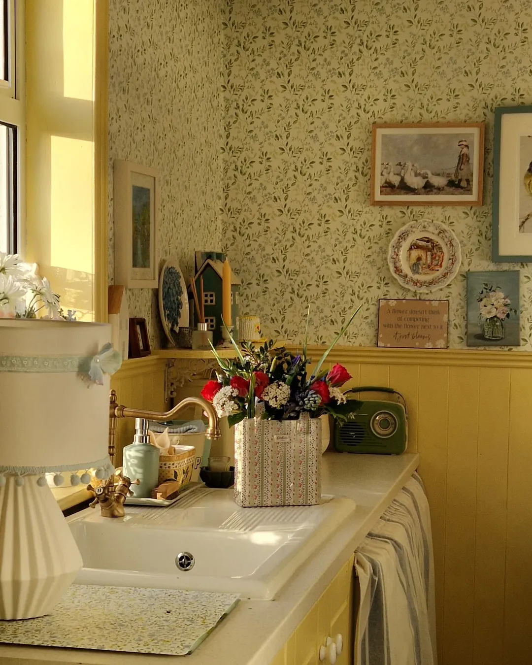 A sunlit vintage kitchen corner featuring cottagecore decor—a sink, floral wallpaper, framed pictures, a vase of artificial flowers, and a green retro radio grace the countertop for cozy Cottagecore kitchen charm.