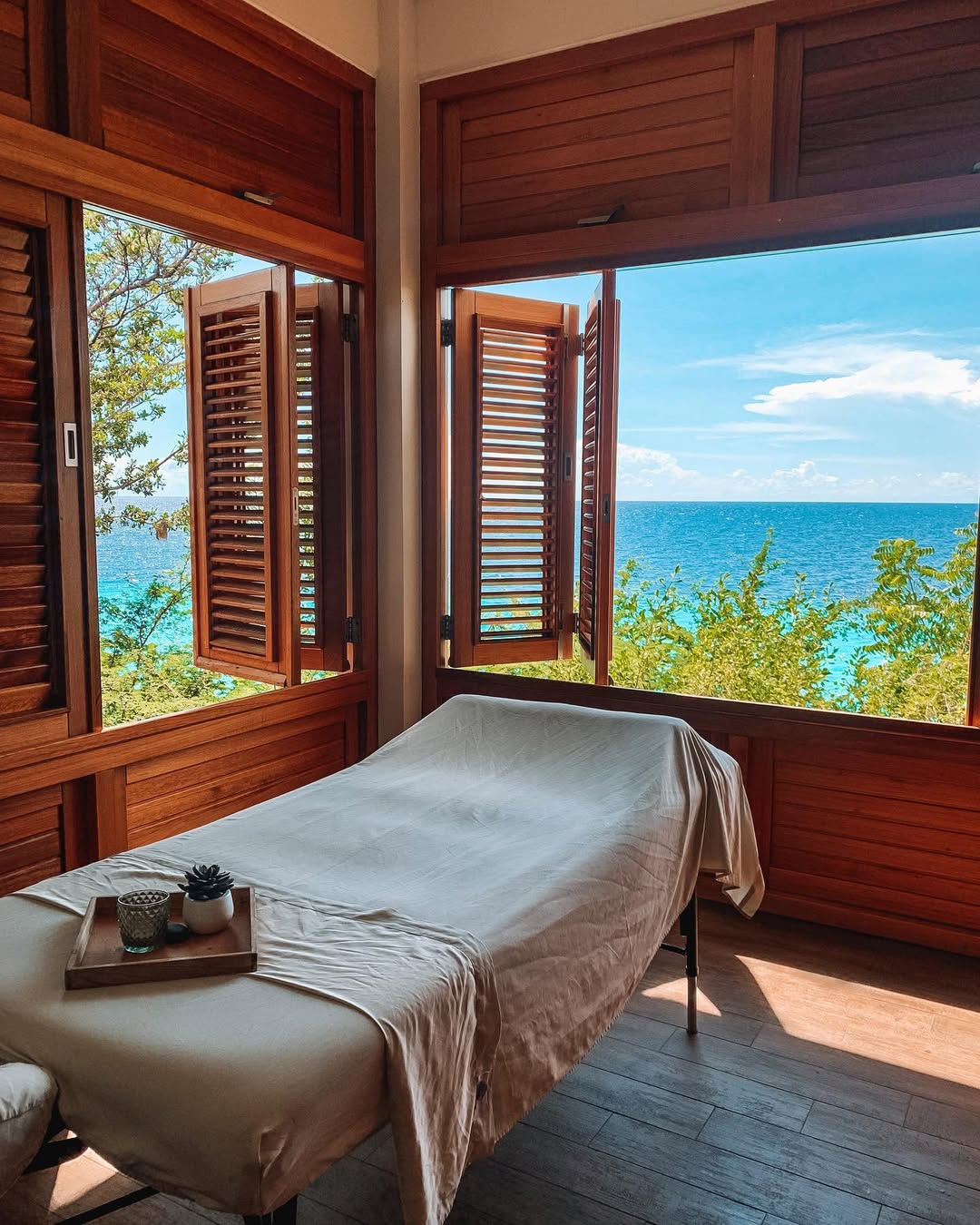 A massage table with a beige sheet is set near large open windows with a view of the ocean and blue sky, offering inspiring massage room ideas. A tray with small items rests on the table.