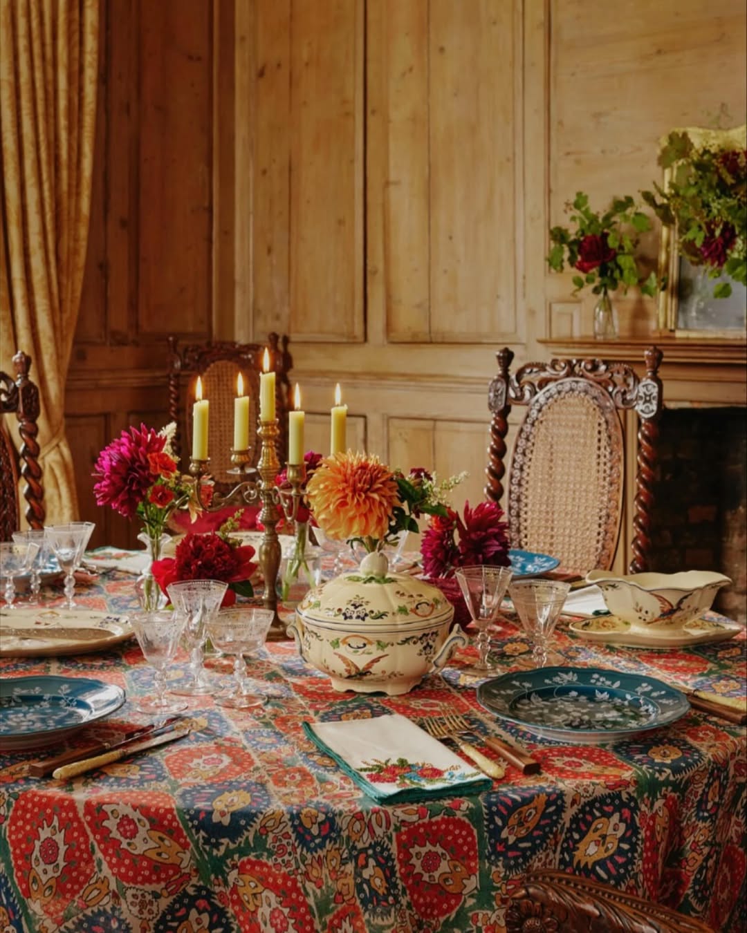 A dining table set with patterned dishes, a floral tablecloth, elegant table decor including a candelabra with lit candles, and vases of flowers, in a wood-paneled room with ornate chairs for inspiring table setting ideas.
