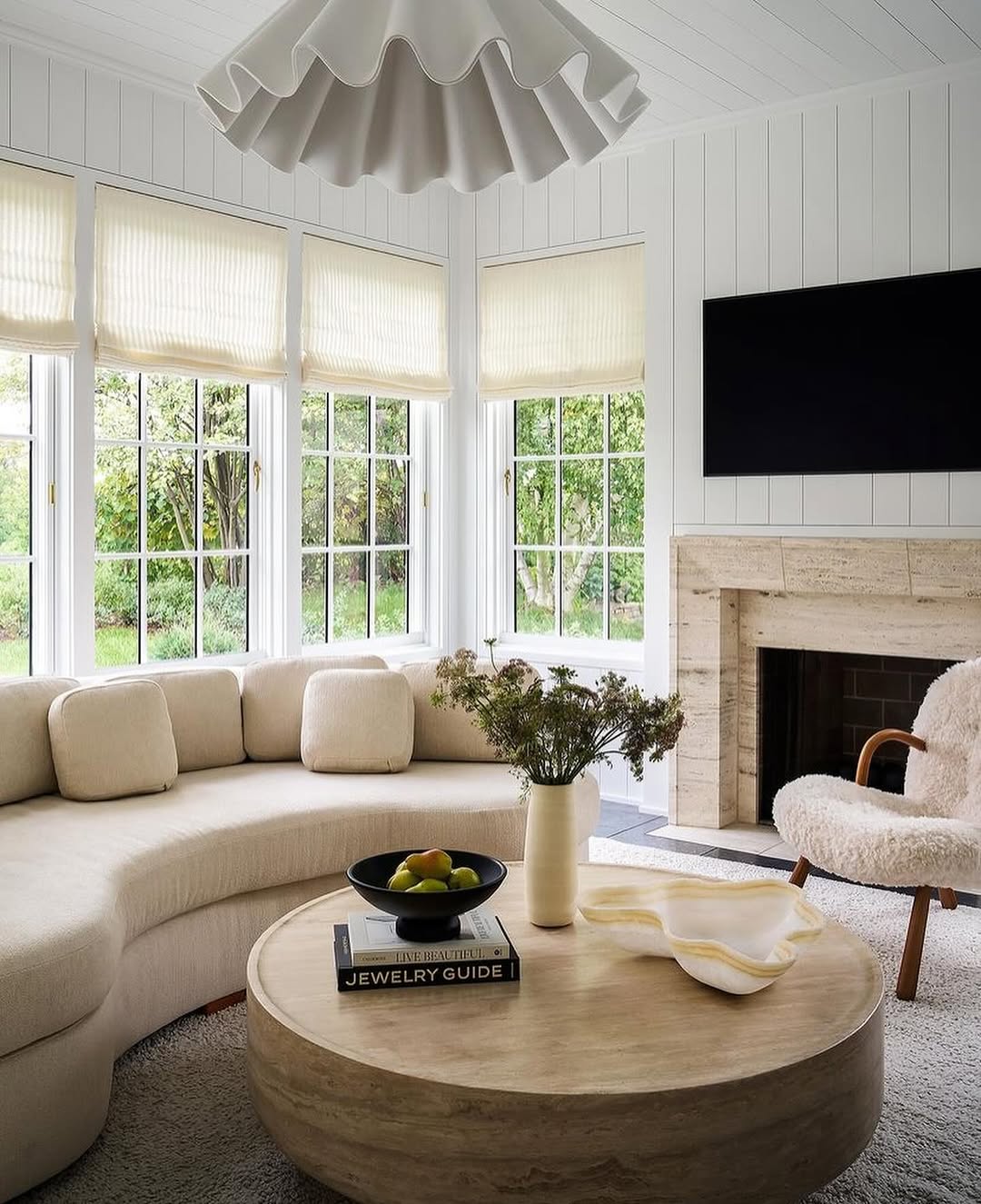 A modern living room featuring a curved cream sofa, round wooden coffee table, fireplace, wall-mounted TV, and large windows offers fresh living room ideas for an airy, light-filled space.