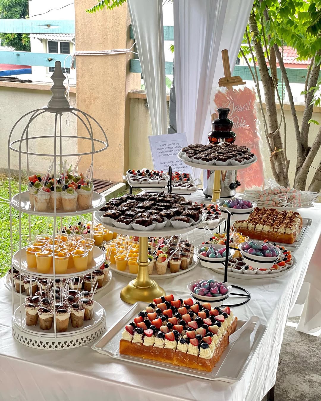 A dessert table set outdoors with occasion desserts like assorted pastries, cakes, fruit tarts, brownies, mousse cups, and chocolate-dipped treats arranged on stands and trays—perfect for inspiring pretty dessert table ideas.