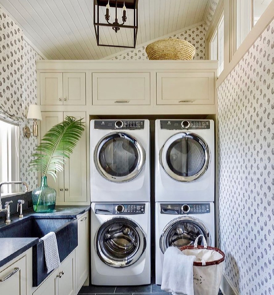 A small laundry room with two stacked washers and dryers features a farmhouse sink, patterned wallpaper, and a basket on top of the cabinets. A green leaf in a vase adds a touch of freshness to the cozy space.