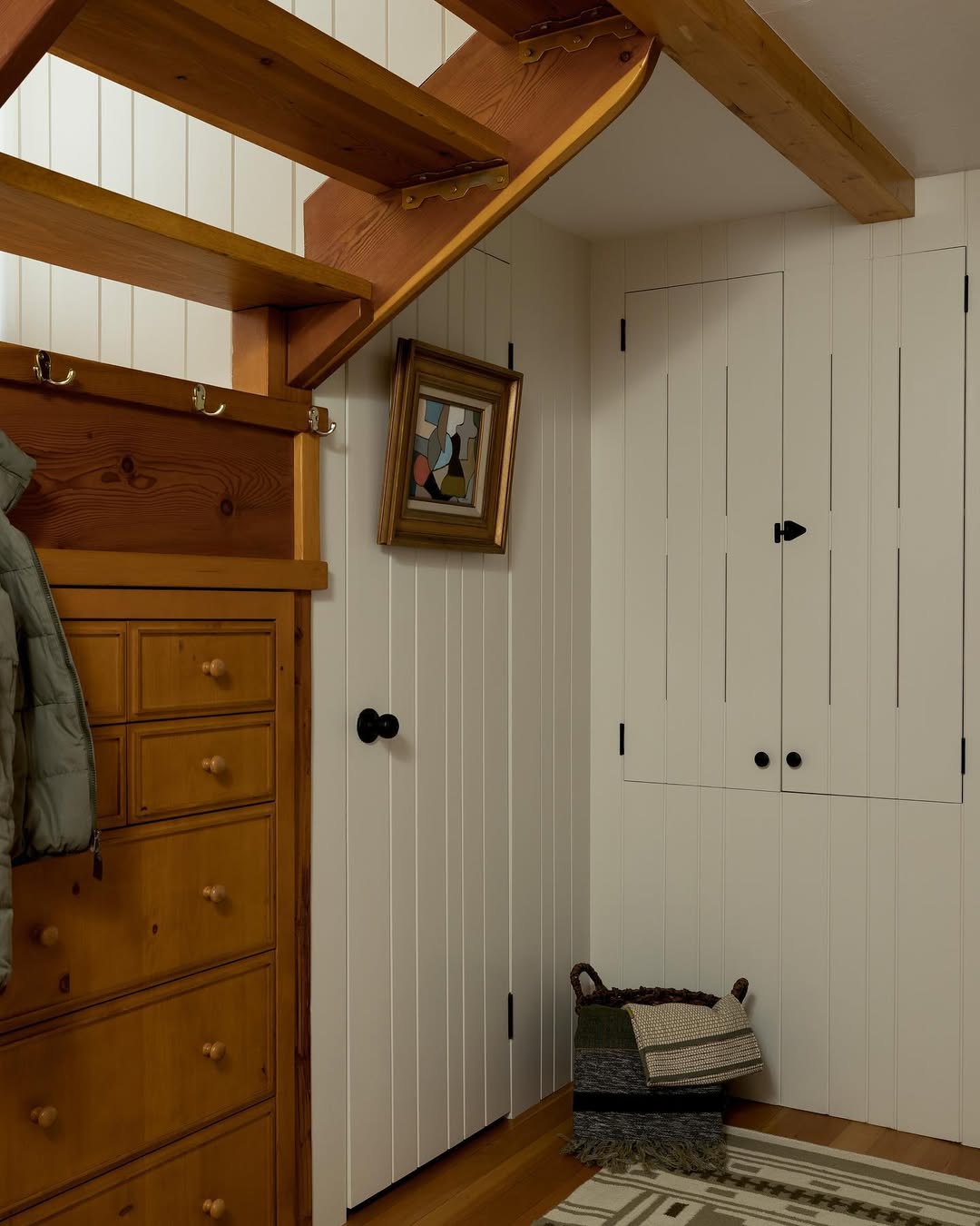 A small entryway features wood stairs, a pine dresser, white paneled walls with under stairs storage doors, a coat hook, a basket with blankets, and a framed painting.