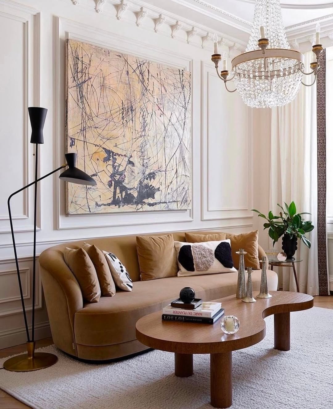 A modern, chic living room featuring a curved tan sofa, abstract wall art, a wooden coffee table with decor, a crystal chandelier, black floor lamp, and potted plant—perfect inspiration for those seeking fresh living room ideas.
