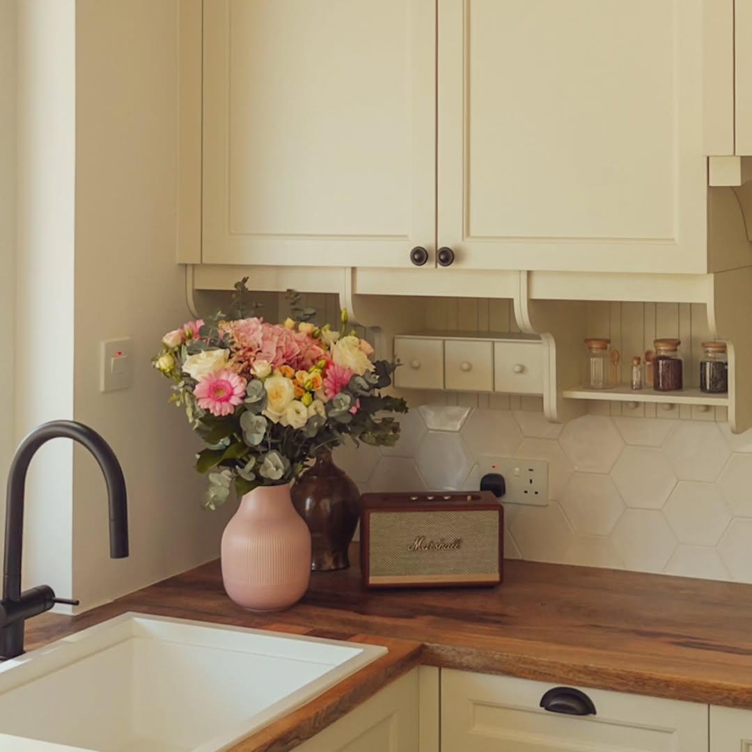A kitchen counter with a pink vase of colorful flowers, a retro-style radio, small jars on a shelf, and a black faucet by a white sink—perfect inspiration for cottagecore kitchen ideas.