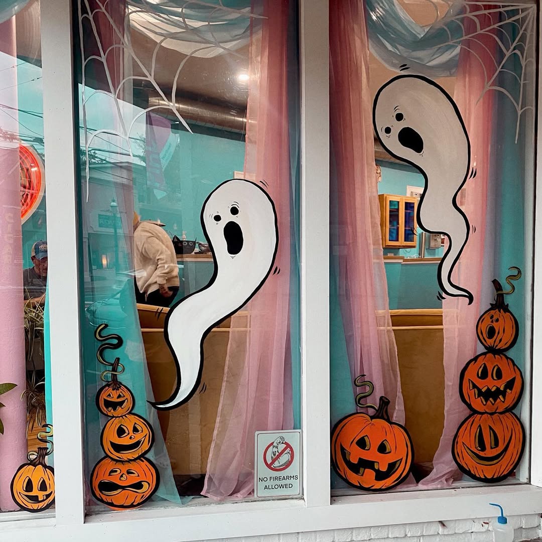 Window decorated with painted cartoon ghosts, pumpkins, spider webs, and pink curtains; a