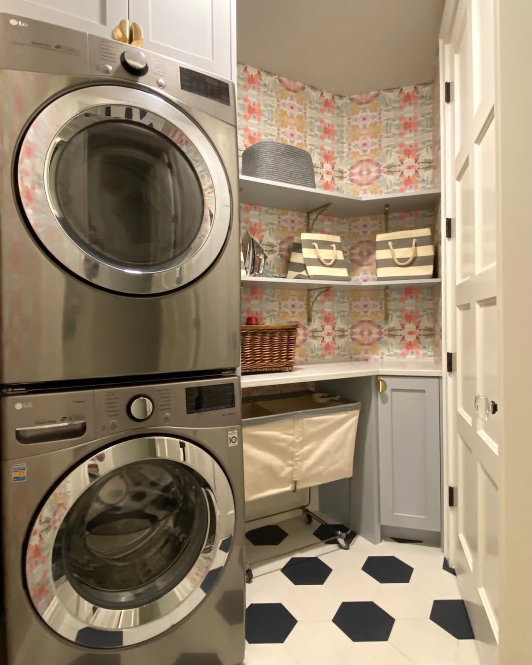 A compact laundry room features a stacked washer and dryer, patterned wallpaper, hexagonal tile floor, shelves with baskets, clever laundry room cabinets, and a rolling laundry hamper.