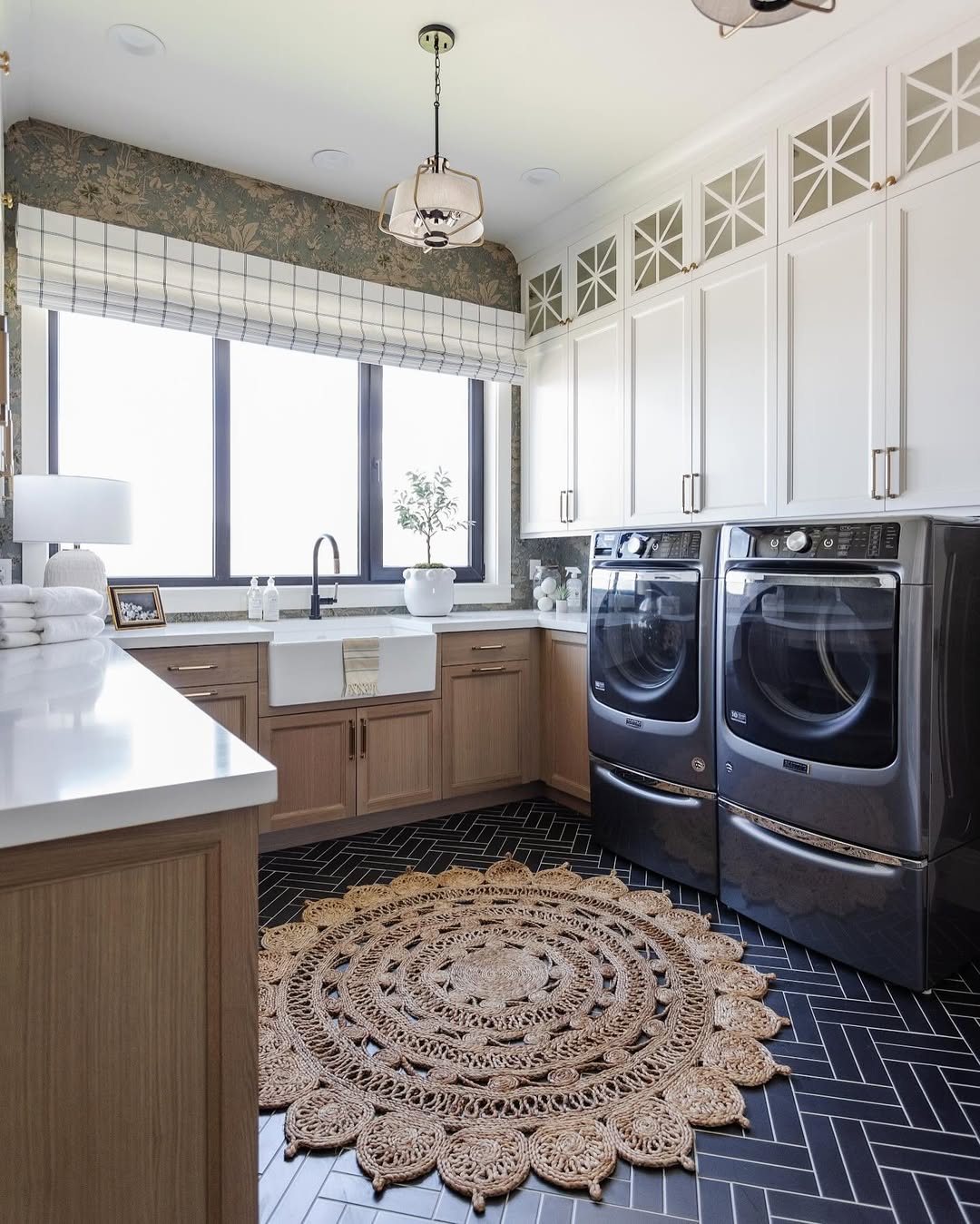 Modern laundry room with white and wood Laundry Room Cabinets, black washer and dryer, farmhouse sink, large window, and a round woven rug on a dark herringbone tile floor.