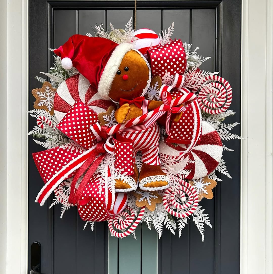 A festive wreath with a gingerbread figure in a Santa hat, red and white ribbons, candy cane decorations, and white foliage on a dark door—perfect for adding Gingerbread Christmas decor to your list of Christmas decor ideas.